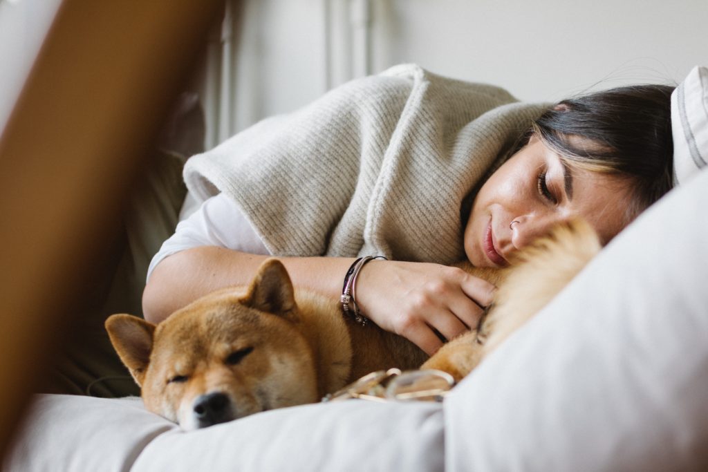 Woman with dark hair takes a nap with Shiba Inu while resting on a couch and wearing a wool cream colored shawl
